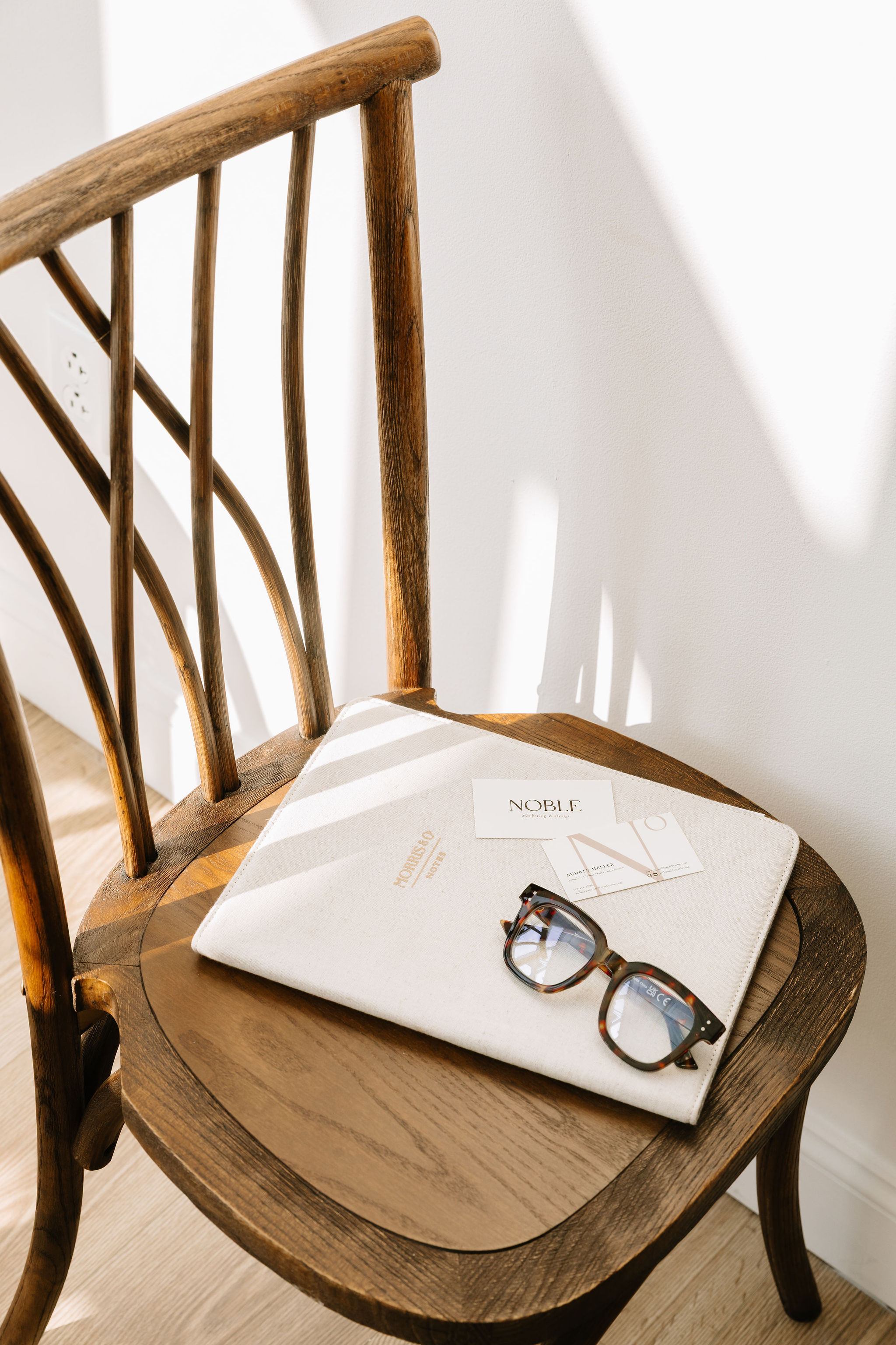 Noble business cards, portfolio sleeve, and glasses on a wooden chair in natural light