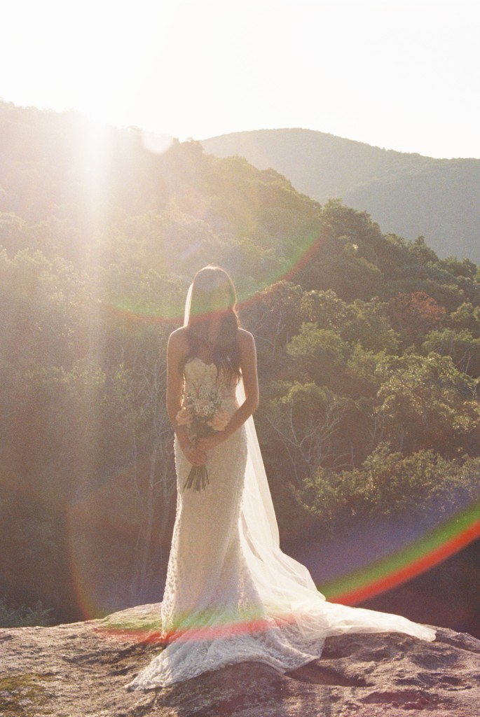 Bride in a lace gown standing on a rock with a bouquet, backlit by sun flare over a forested mountain