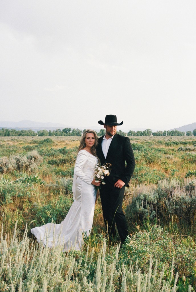 Bride in a white gown and groom in a black suit and cowboy hat standing together in a sagebrush field