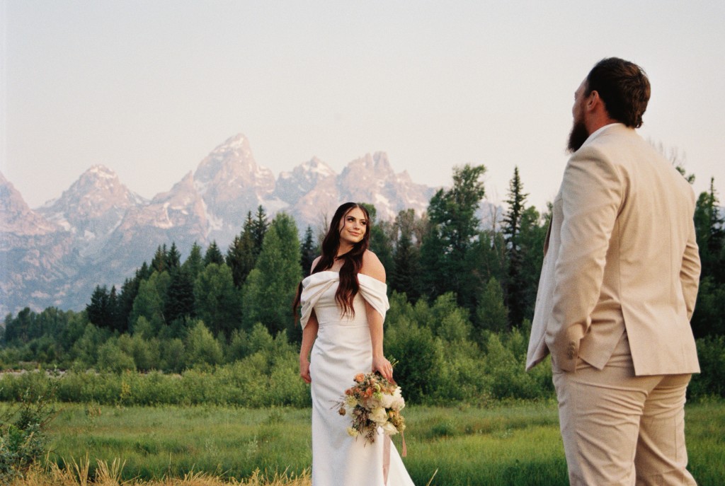 Bride in an off-shoulder gown holding a bouquet in a meadow, with a groom in a light suit and snow-capped mountains behind them