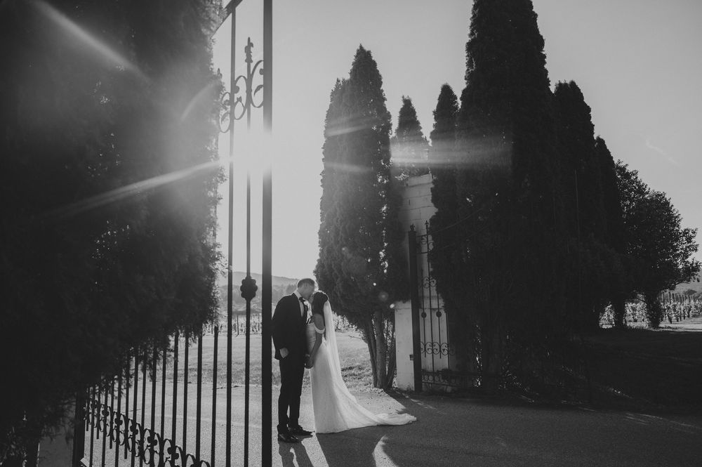 Black-and-white photograph of a bride and groom kissing beside a wrought-iron gate and tall cypress trees