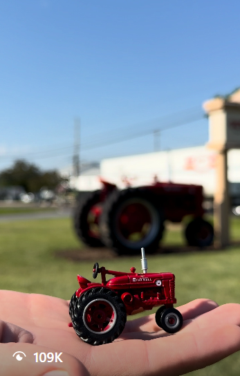 A small Farmall M toy tractor held in a palm with a full-size red tractor behind it
