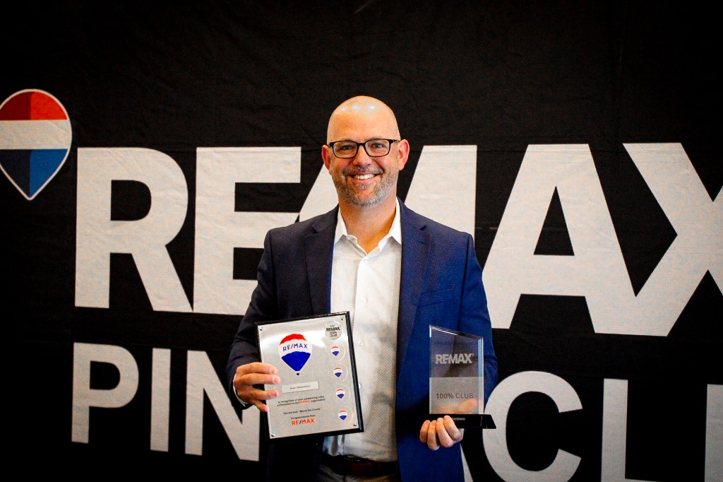 RE/MAX Pinnacle agent holding a 100% Club trophy and Chairman's Club plaque