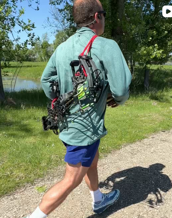Runner on a gravel path with a compound bow secured across the back by a red Vsling