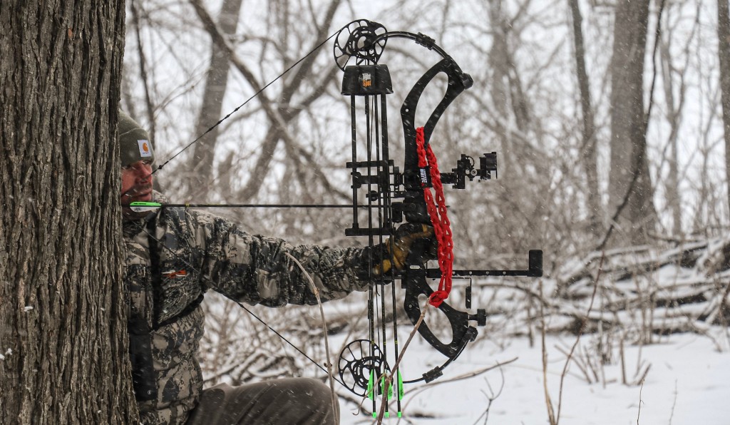 Hunter in winter camouflage aiming a compound bow from behind a tree, with a bright red Vsling on the riser